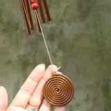 Someone holding the spiral clapper from the Seven Stones Bronze Woodstock Chakra Chime in front of a concrete wall.