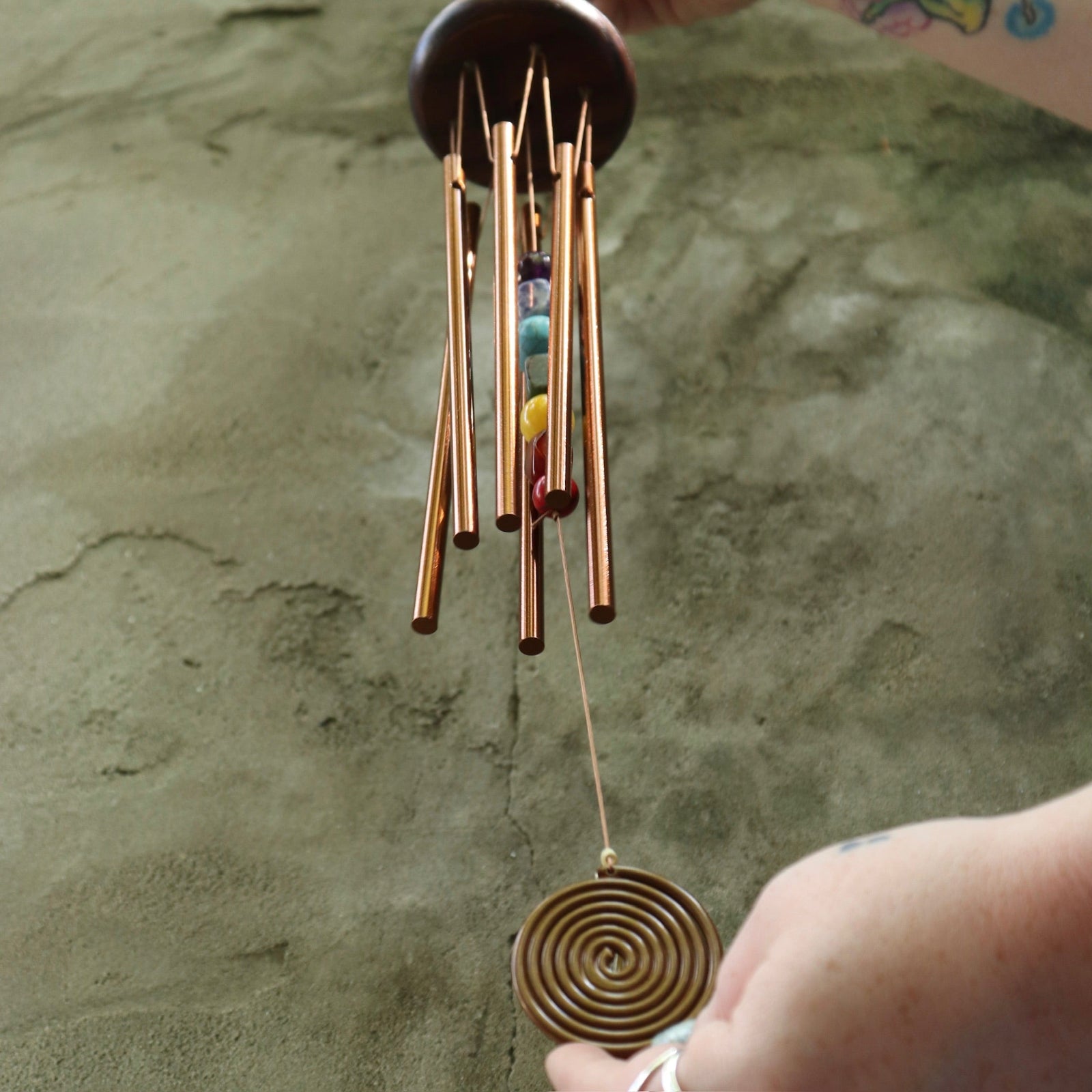Someone holding the Seven Stones Bronze Woodstock Chakra Chime in front of a concrete wall.
