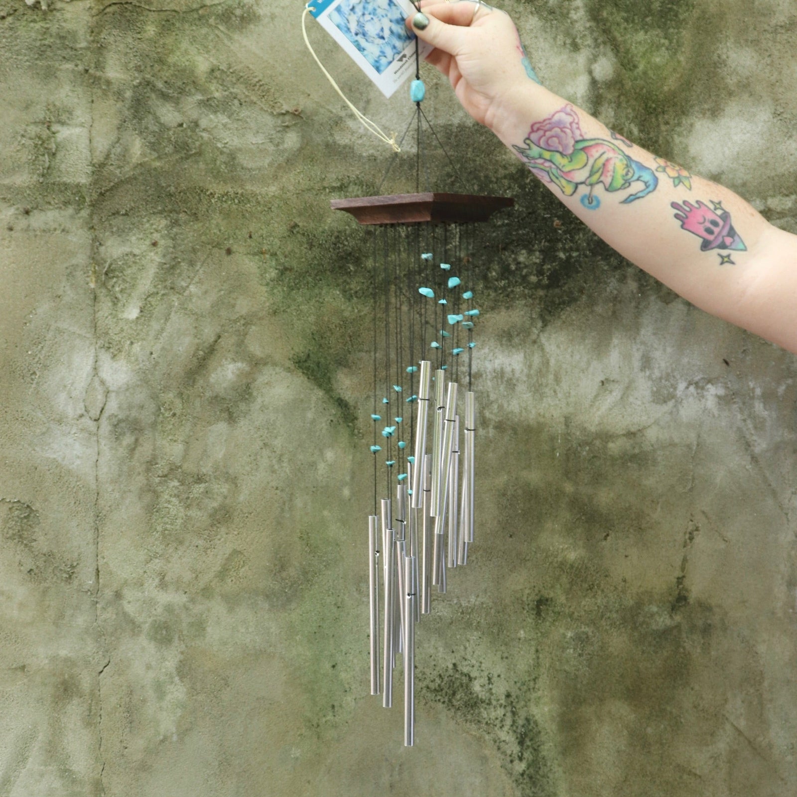 Someone holding the Turquoise Mystic Spiral Woodstock Wind Chime in front of a concrete wall.