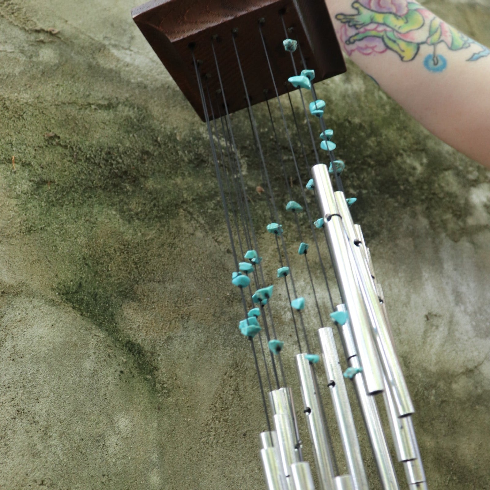 Someone holding the Turquoise Mystic Spiral Woodstock Wind Chime in front of a concrete wall.