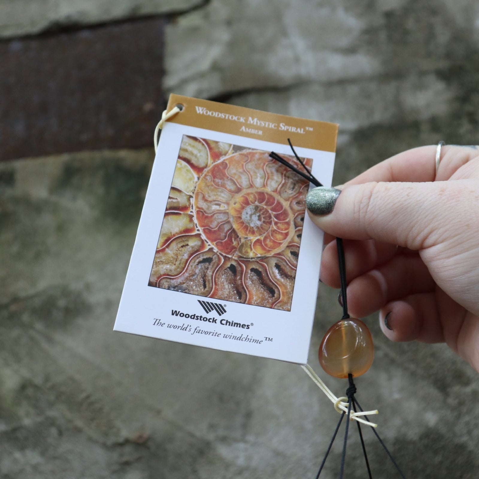 An up-close image of someone holding the tag of the Amber Mystic Spiral Woodstock Wind Chime in front of a concrete wall.