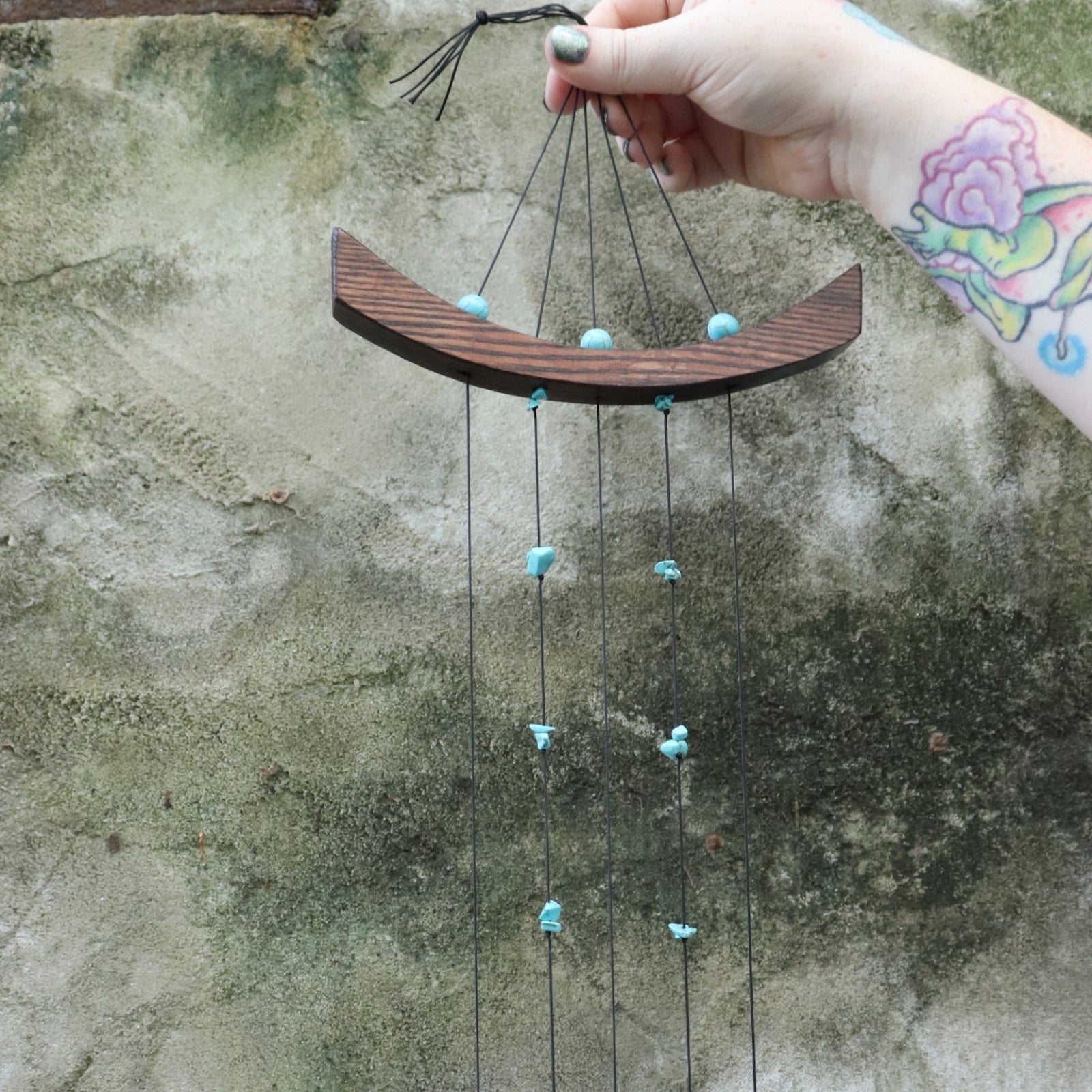 An up-close image of someone holding the Small Turquoise Chi Energy Wind Chime in front of a cement wall.