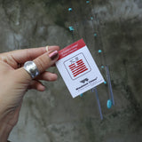 An up-close image of someone holding the tag of the Small Turquoise Chi Energy Wind Chime in front of a cement wall.