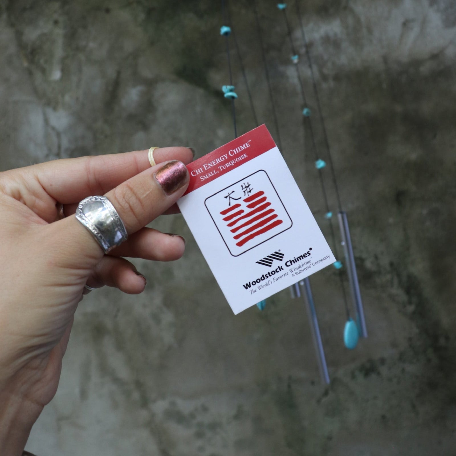 An up-close image of someone holding the tag of the Small Turquoise Chi Energy Wind Chime in front of a cement wall.