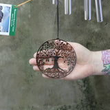 An up-close image of someone holding the Mirror Tree of Life Woodstock Habitats Wind Chime ornament in front of a cement wall.