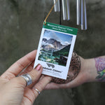 An up-close image of someone holding the tag of the Mirror Tree of Life Woodstock Habitats Wind Chime ornament in front of a cement wall.