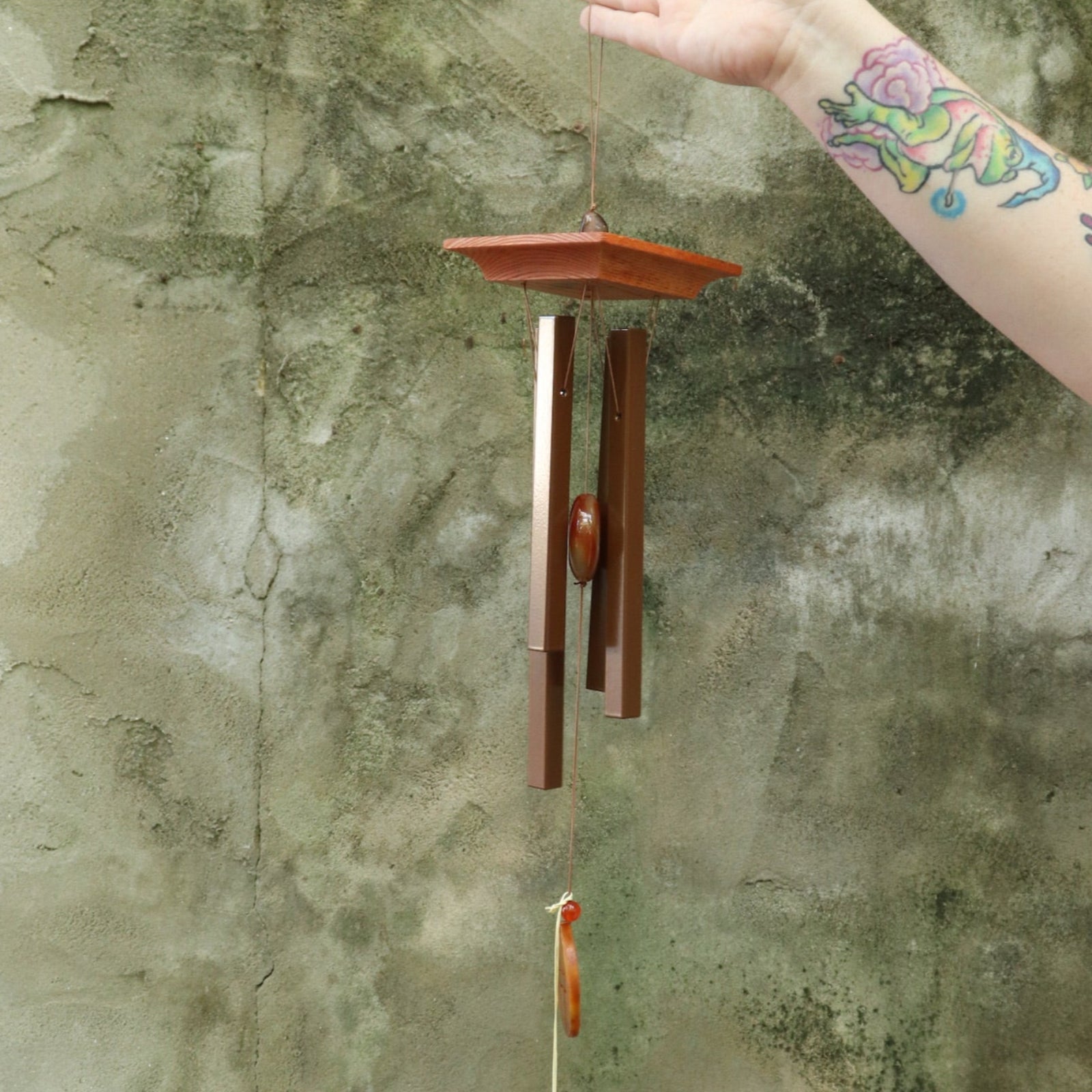 Someone holding the Woodstock Amber Wind Chime in front of a cement wall .