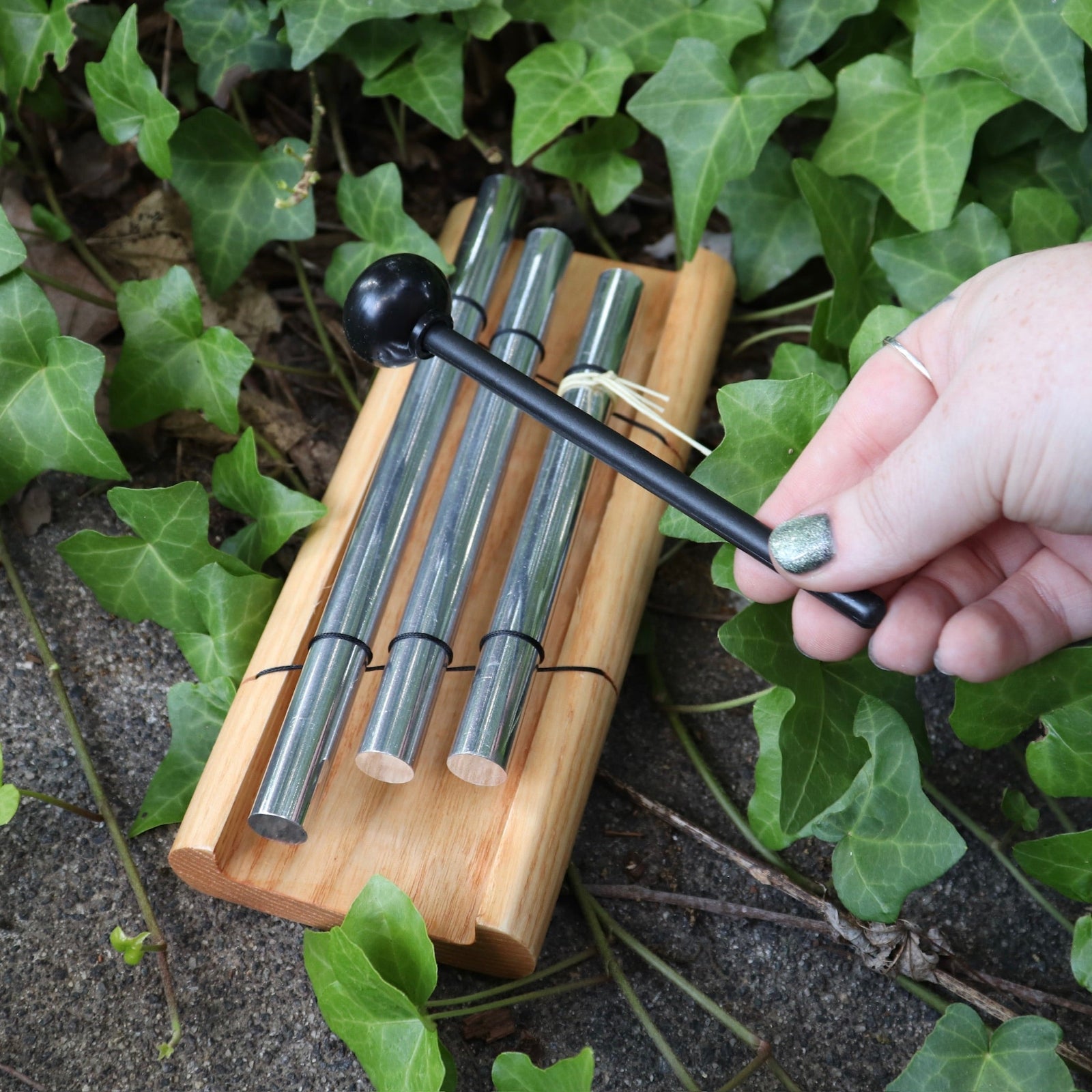 The Woodstock Zenergy Trio Chime on a dark cement background surrounded by ivy, with someone holding the rubber mallet.
