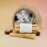 The Palo Santo Smudge Bundle by Bohemian Rêves in front of a cracked geode and surrounded by crystals all on a pink and yellow background.