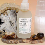 An up close image of the Patchouli Vanilla Sea Salt Texturizing Hair Mist by Bohemian Rêves on an agate slice in front of a cracked geode and a selenite tower all on a pink and yellow background.