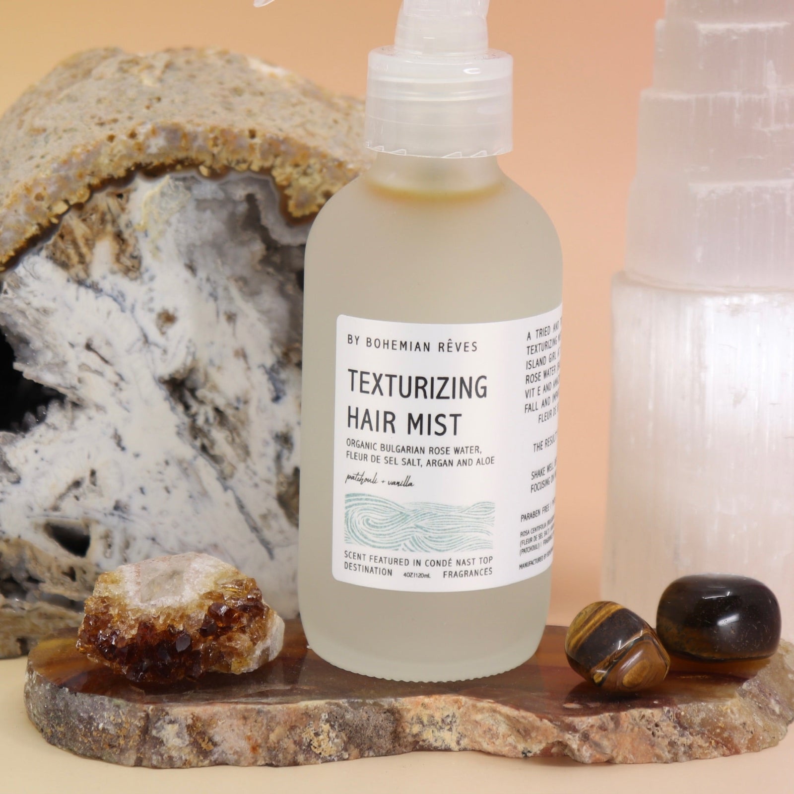 An up close image of the Patchouli Vanilla Sea Salt Texturizing Hair Mist by Bohemian Rêves on an agate slice in front of a cracked geode and a selenite tower all on a pink and yellow background.