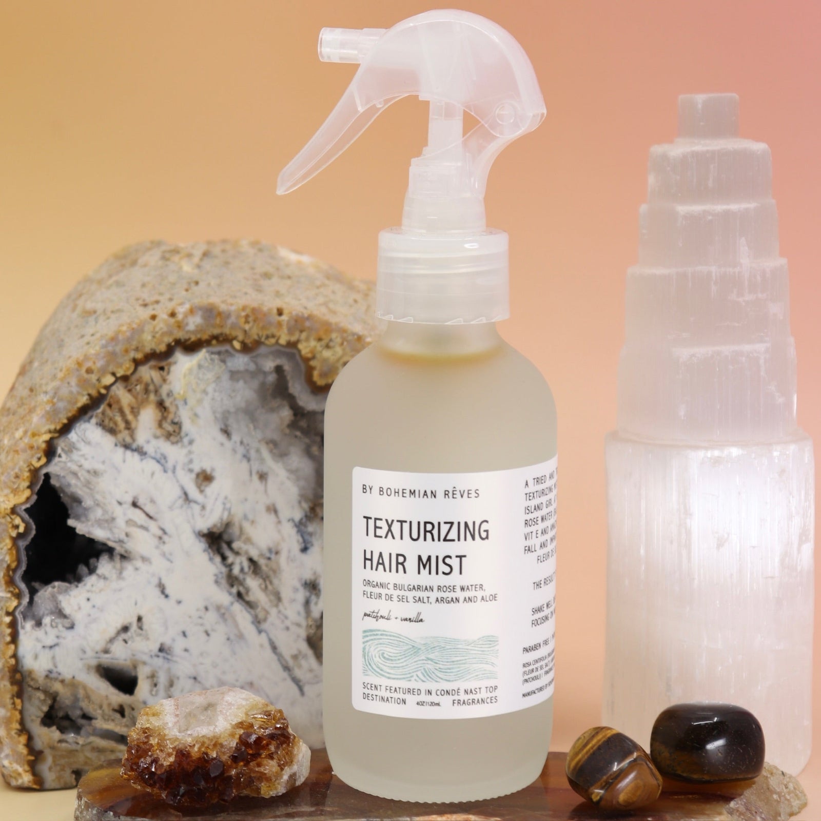 The Patchouli Vanilla Sea Salt Texturizing Hair Mist by Bohemian Rêves on an agate slice in front of a cracked geode and a selenite tower all on a pink and yellow background.
