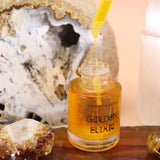 The Anti-Aging Elixir Face Oil from Bohemian Rêves out of its box and on a slice of agate in front of a open geode and selenite tower, all on a pink and yellow background.