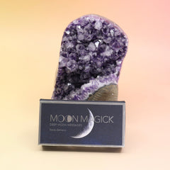 The Moon Magick Affirmation + Divination Cards against an amethyst piece all on a pink and yellow background.