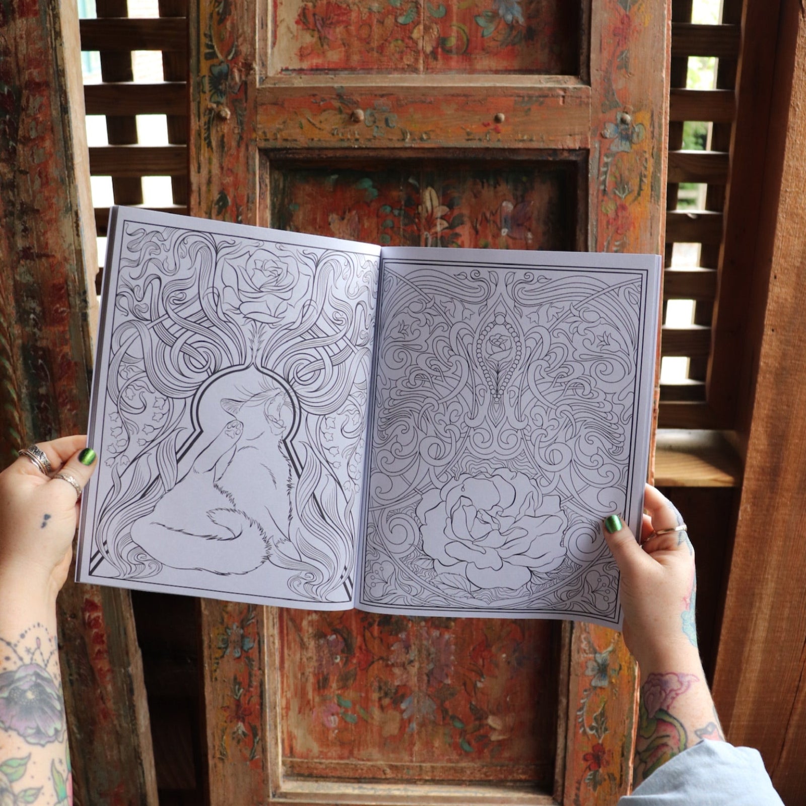 Someone holding the Cats and Roses Coloring Book open in front of a wooden background, showing off the pages.