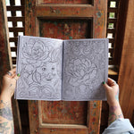 Someone holding the Cats and Roses Coloring Book open in front of a wooden background, showing off the pages.