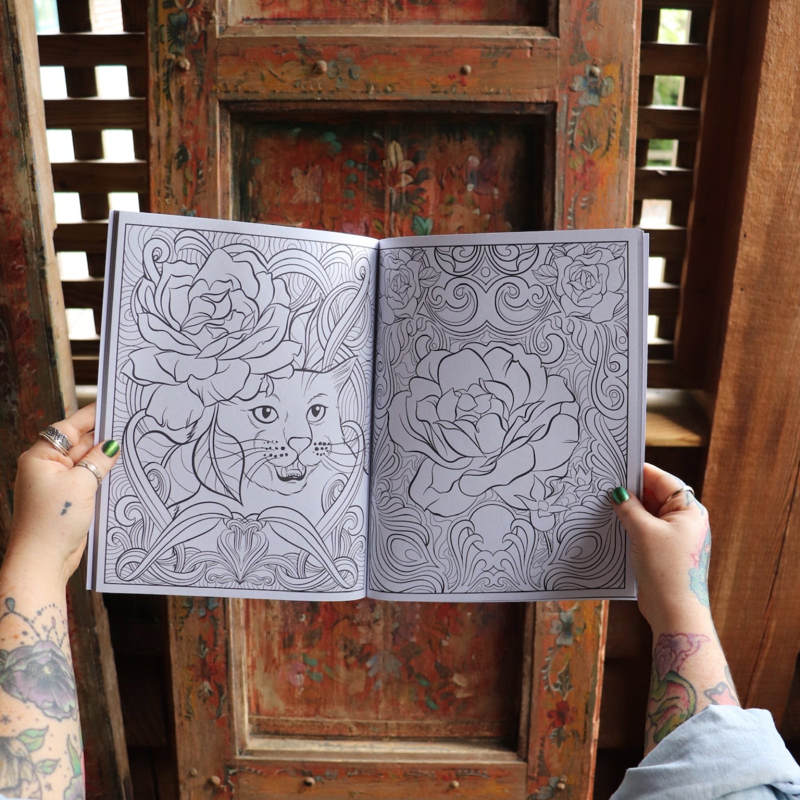 Someone holding the Cats and Roses Coloring Book open in front of a wooden background, showing off the pages.