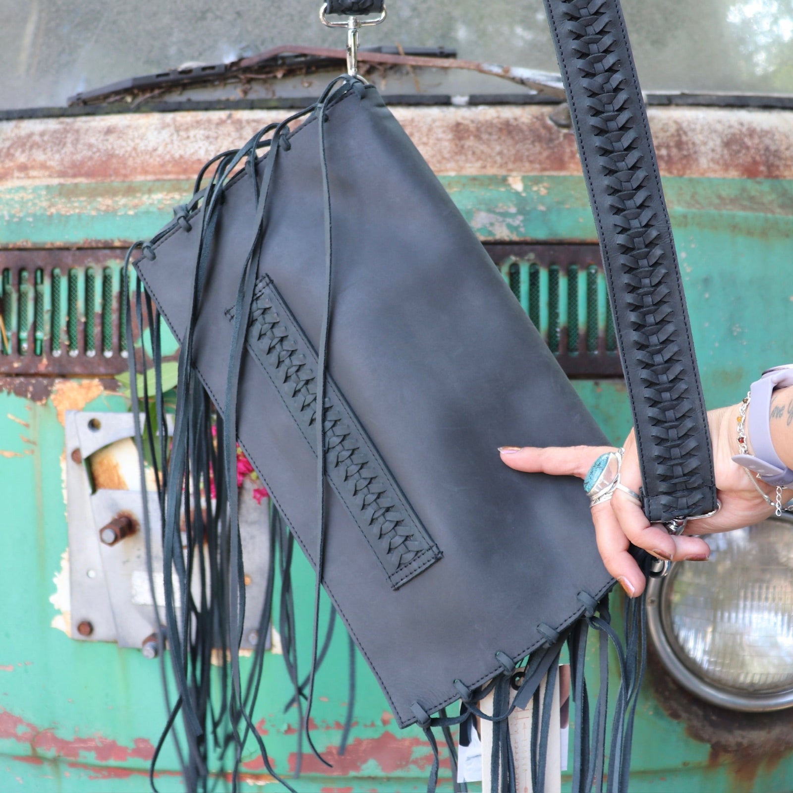 Black leather clutch handbag with fringe details held by a person against a rustic background.