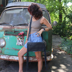 Woman leaning against a vintage green van with a black leather clutch handbag, surrounded by greenery.