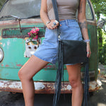 Person wearing a beige top and denim shorts with a black fringed leather clutch bag, standing in front of a vintage green truck.
