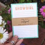 Notepad titled 'The To-Do List of a Showgirl' held by a hand with flowers in the background