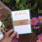 Hand holding a notepad with 'She is a Showgirl' text against a floral background