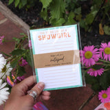Hand holding a notepad with 'She is a Showgirl' text against a floral background