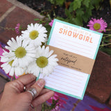 Hand holding a notepad titled 'The To-Do List of a Showgirl' with flowers in the background