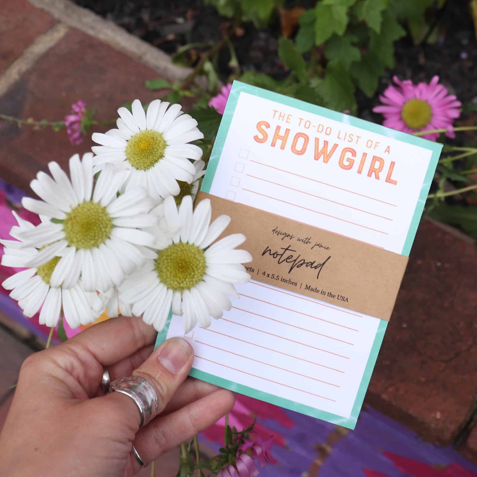 Hand holding a notepad titled 'The To-Do List of a Showgirl' with flowers in the background
