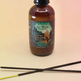 up close image of the litha sea witch botanicals spray with 2 crossed incense sticks on an orange and pink background