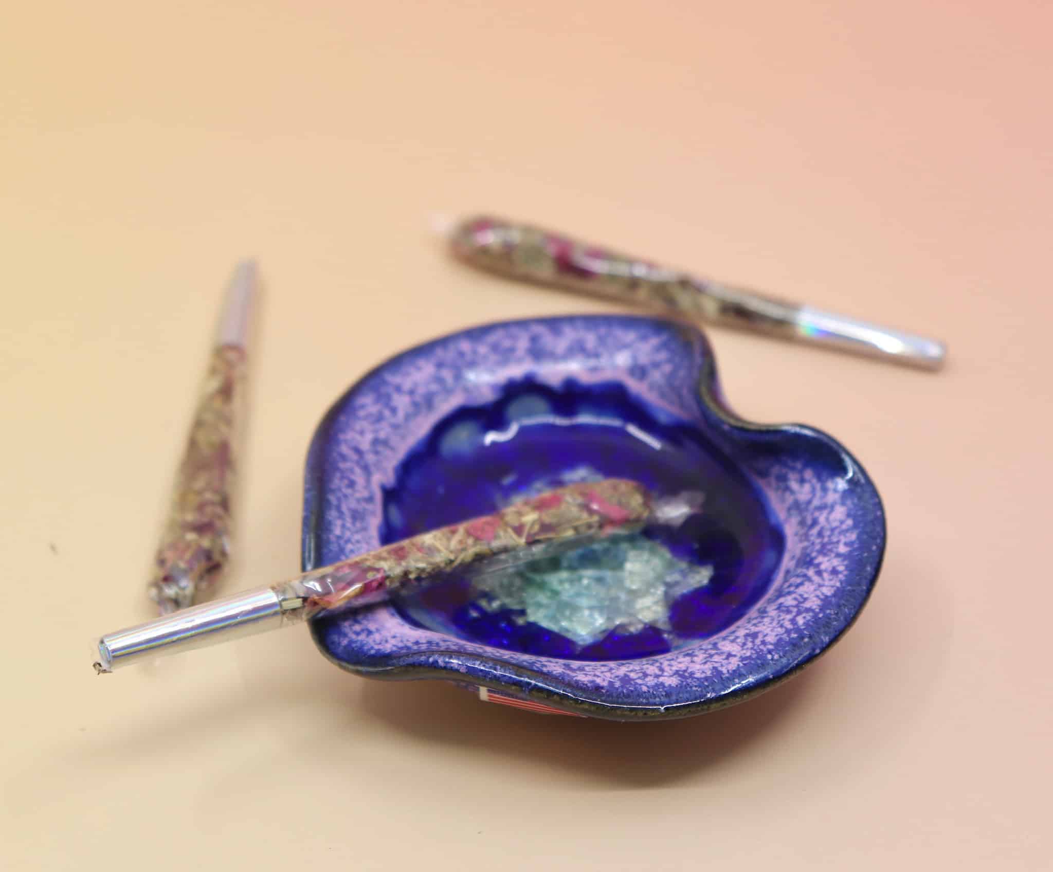 Holographic package of Manifest Magic Rollies with visible herbs inside, surrounded by individual rollies on a soft blush background