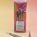 Holographic package of Manifest Magic Rollies with visible herbs inside, surrounded by individual rollies on a soft blush background