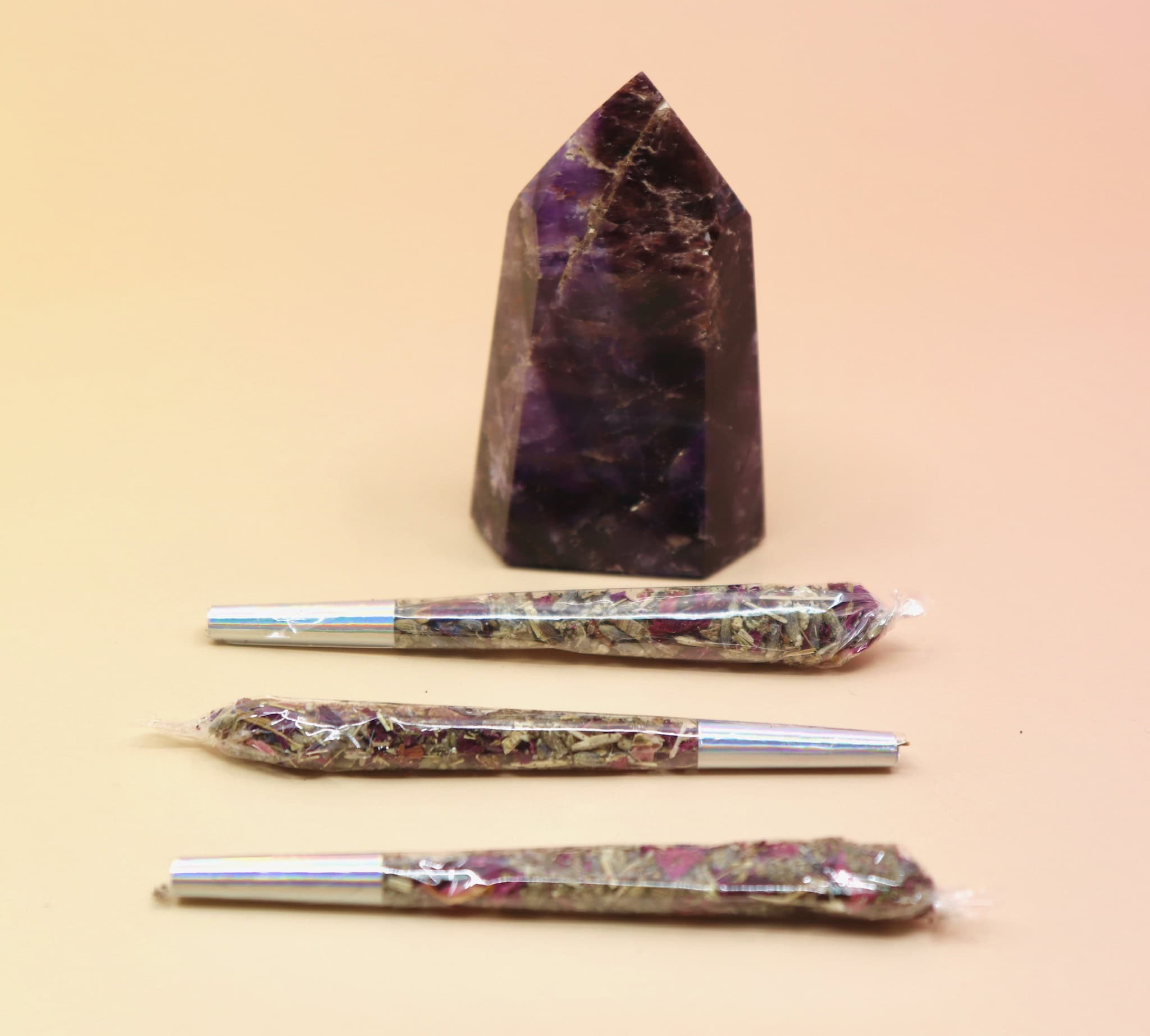 Holographic package of Manifest Magic Rollies with visible herbs inside, surrounded by individual rollies on a soft blush background