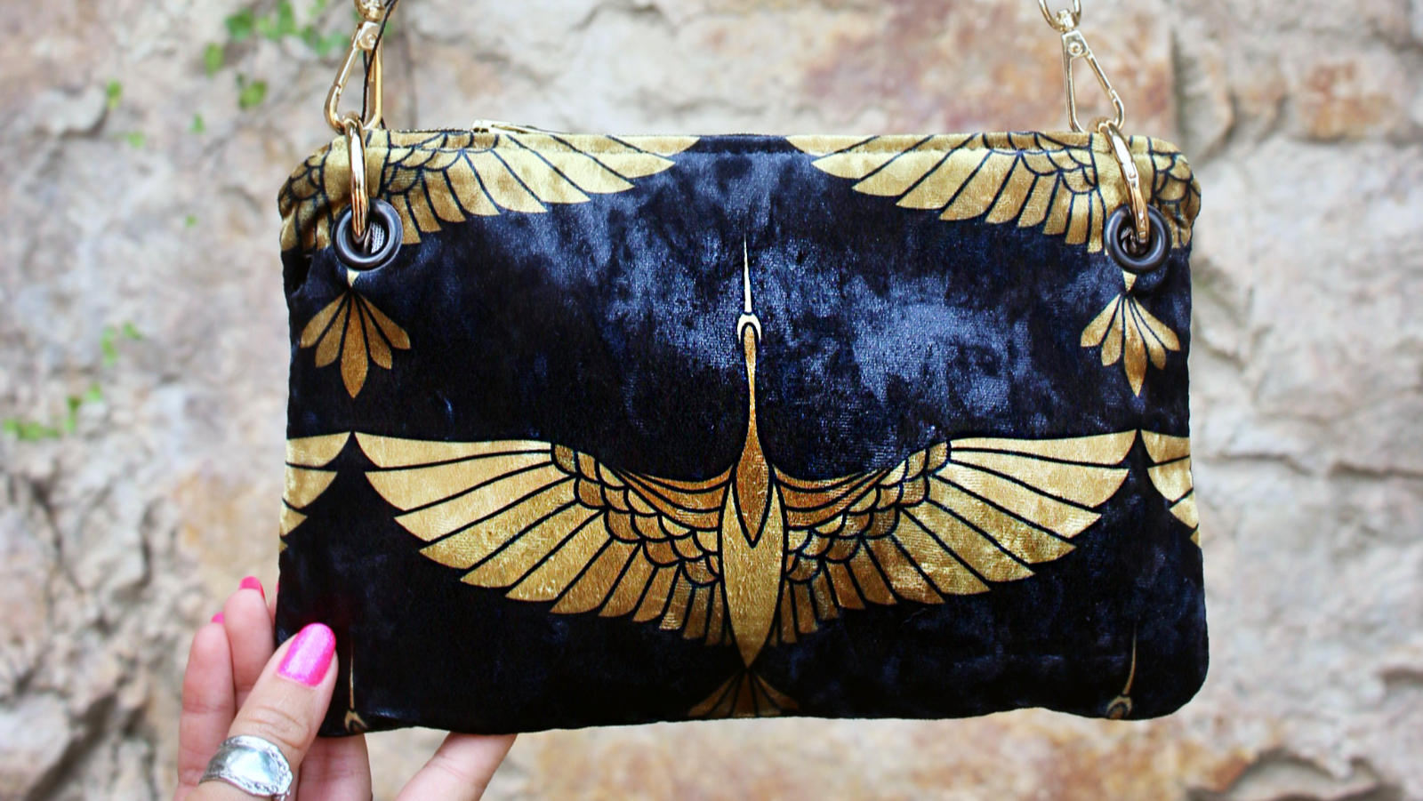 Handbag with gold wing design held against a stone wall background