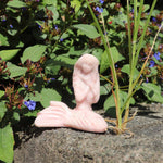 Pink opal crystal stone mermaid sculpture of a reclining figure on a rock with greenery and flowers in the background