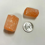 two orange selenite cylinder tumbles next to a U.S. quarter for size comparison