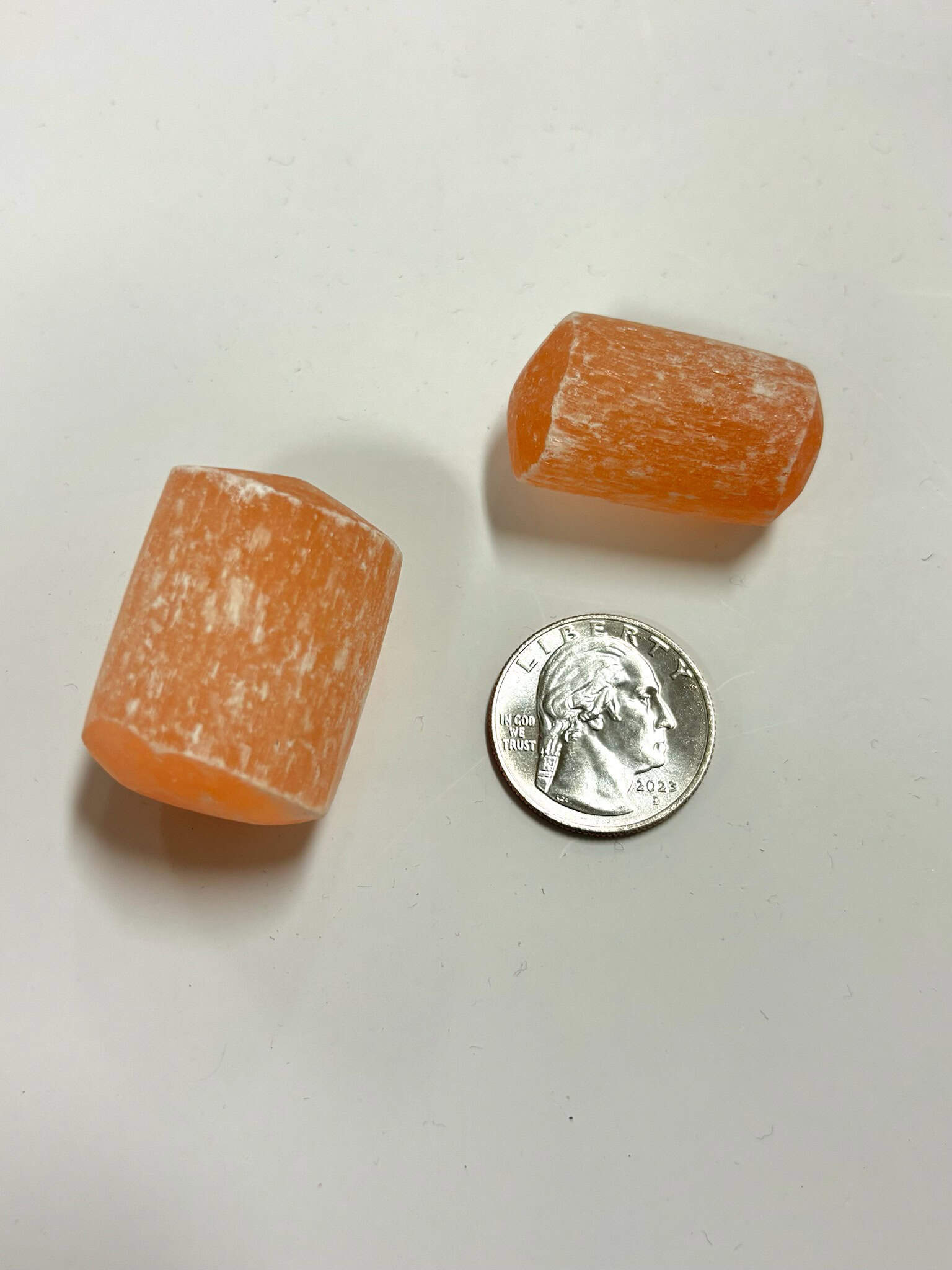 two orange selenite cylinder tumbles next to a U.S. quarter for size comparison