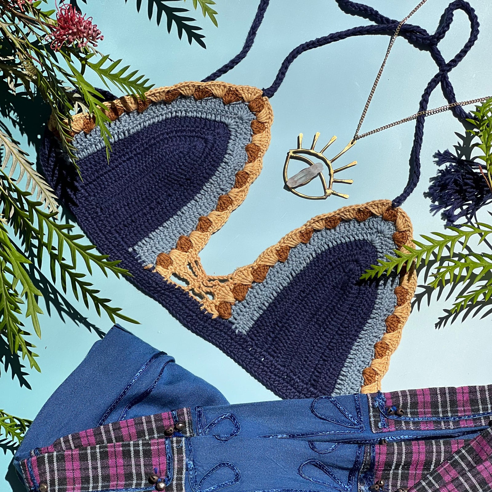The navy Festive Crochet Top: One Size on a blue background with blue pants, an evil eye necklace, and green stems surrounding the top.