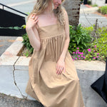 A woman wearing the khaki Solid Color Pleats Suspender Acetate Fiber Free Size Dress while sitting on a white ledge.