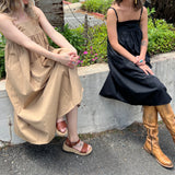 Two women wearing the Solid Color Pleats Suspender Acetate Fiber Free Size Dresses in black and khaki while sitting on a white ledge in front of greenery.