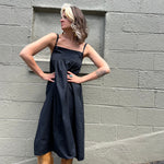A woman wearing the black Solid Color Pleats Suspender Acetate Fiber Free Size Dress in front of a white brick wall.