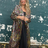 A woman wearing the Rainbow Rage Sequin Cocoon Jacket with a black dress in front of a blue distressed wall.