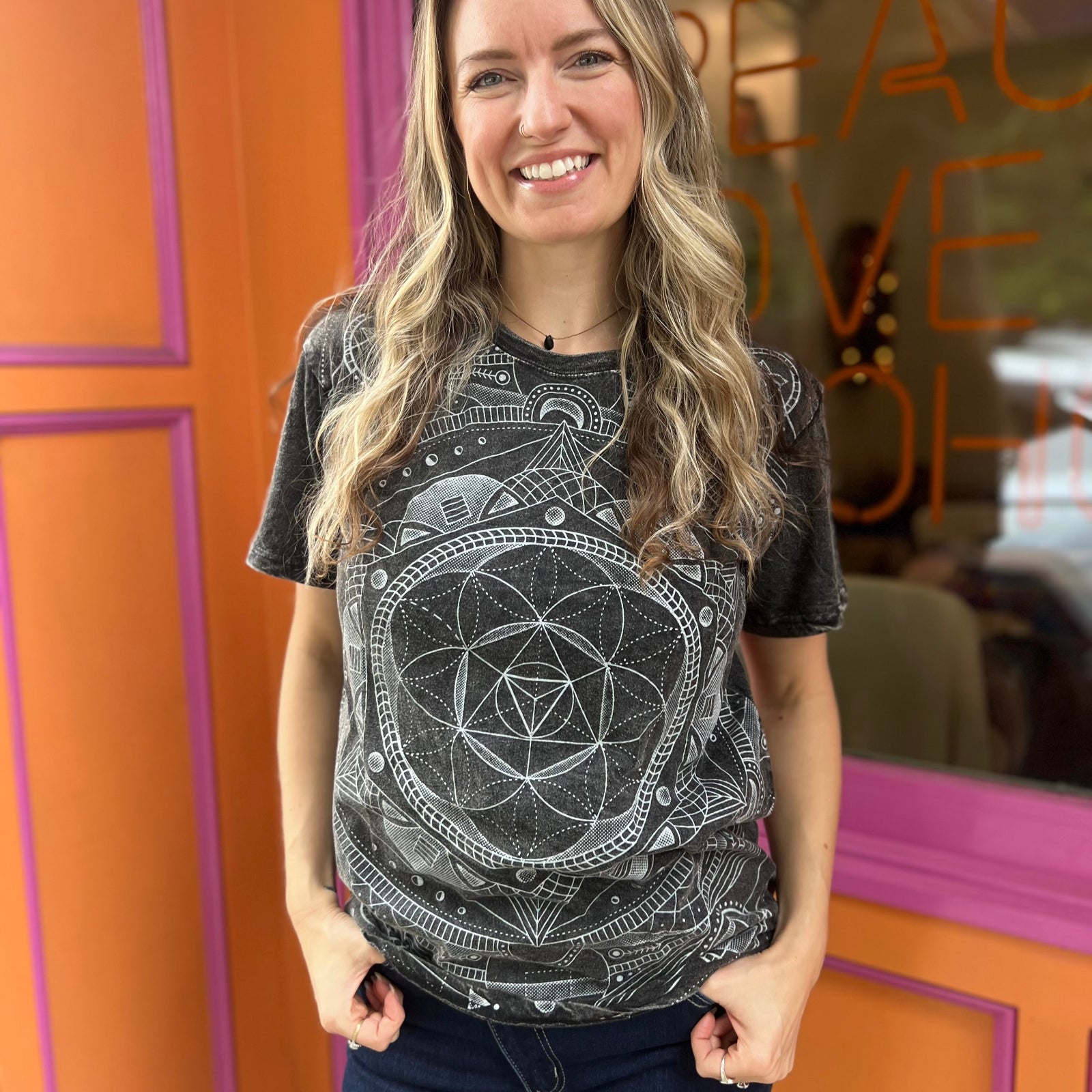 A woman wearing the Gray Mandala Stonewashed Cotton T-shirt with jeans in front an orange and pink wall.
