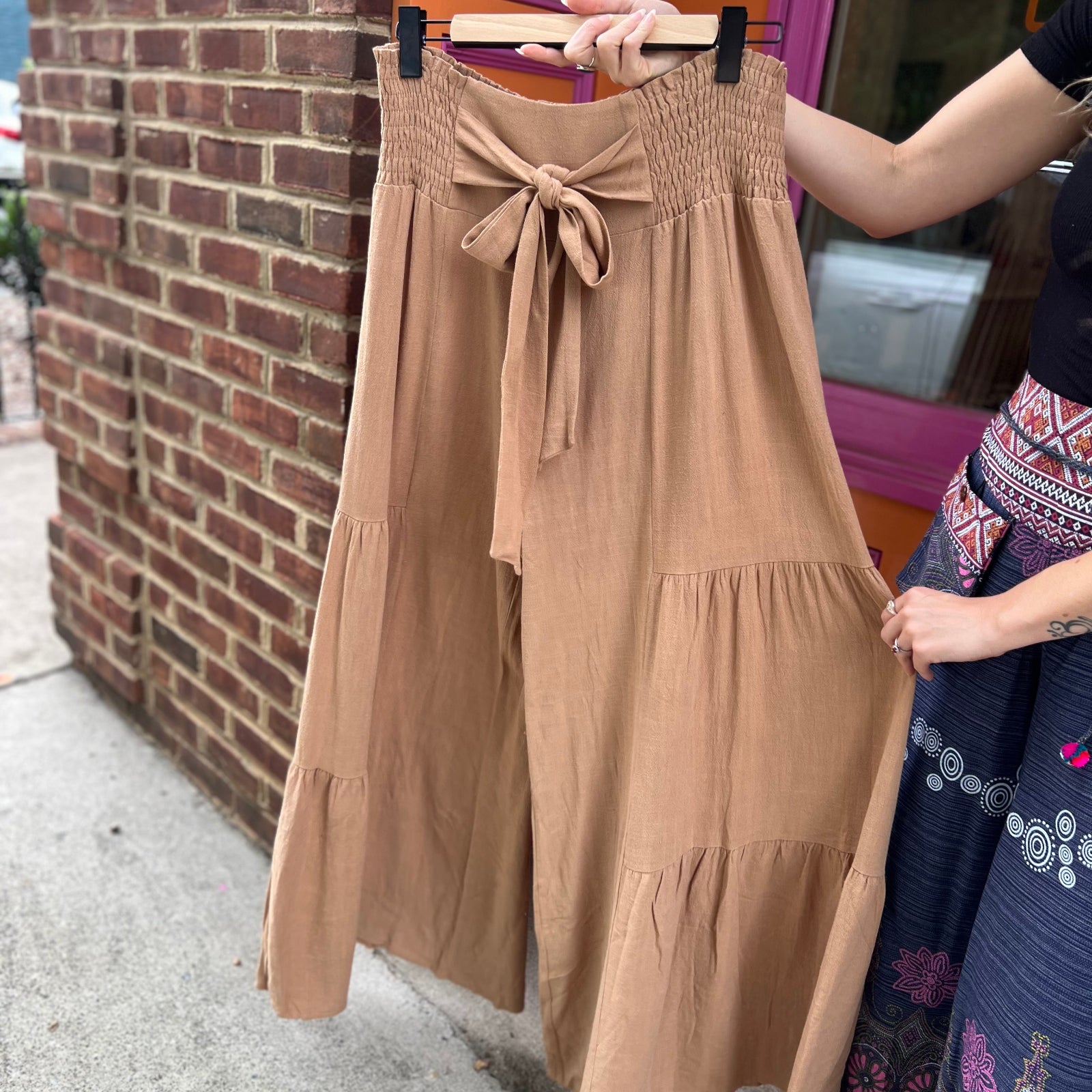 Someone holding up the Plus Smocking Waist Tier Wide Pants in front of a brick wall.