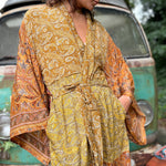 A woman the Hippie Golden Patchwork Kimono Robe Duster with a white bralette and red velvet bellbottoms in front of a vintage Volkswagen bus.