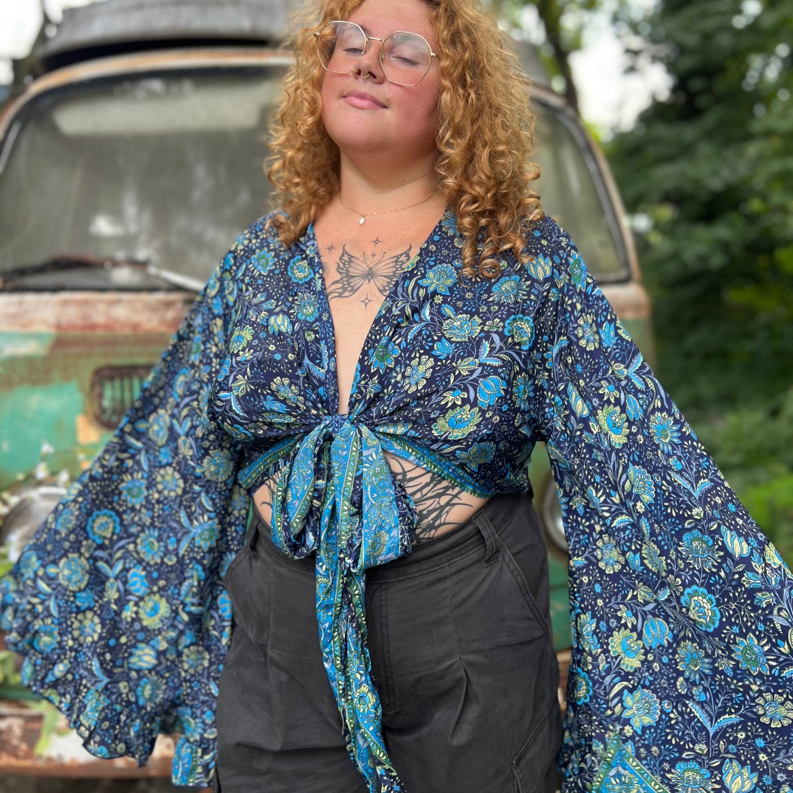 A woman wearing the blue Hippie Bohemian Bell Sleeve Wrap Top with black pants in front of a rusted vintage Volkswagen bus.