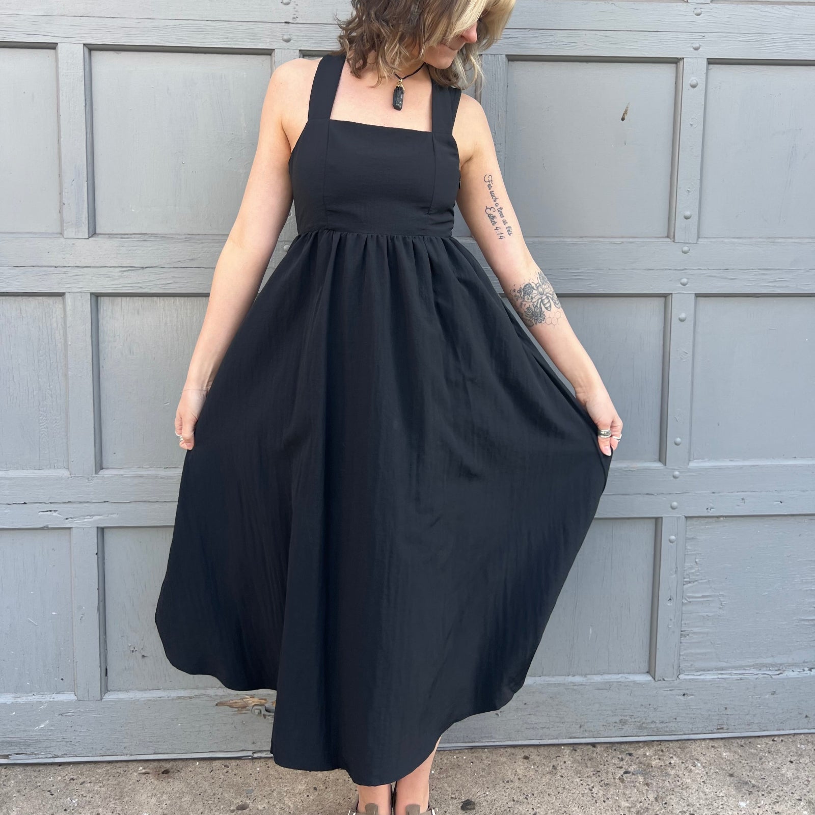 A woman wearing the Black Woven Polyester Nylon Back Cross Strap Midi Dress with sandals and a black crystal necklace.
