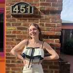 A woman wearing the Contrast Cotton Flare Bow Top with white jeans in front of a brick wall.