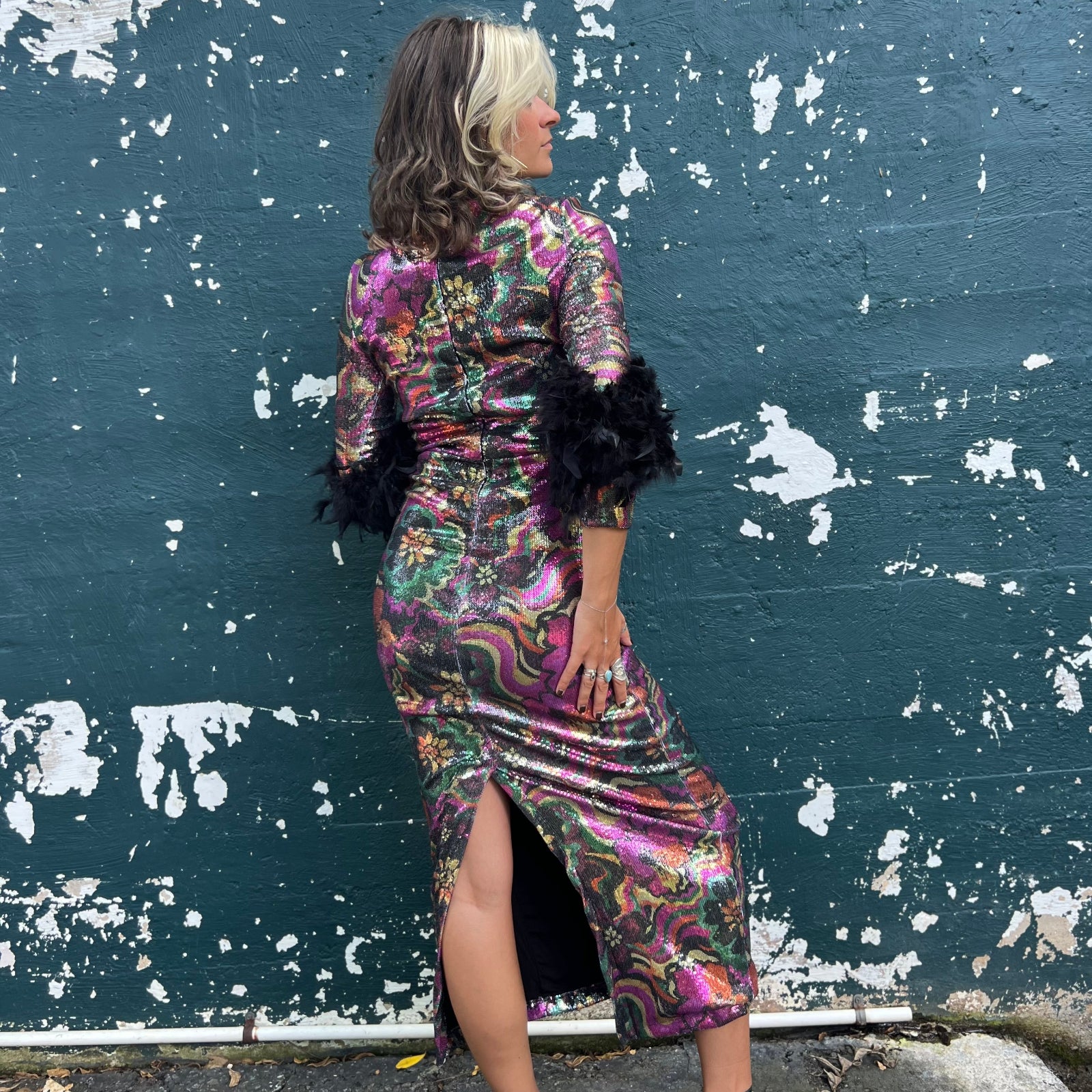 A woman wearing the Catroux dress in Hysteria with a pair of heels in front of a dark teal wall.