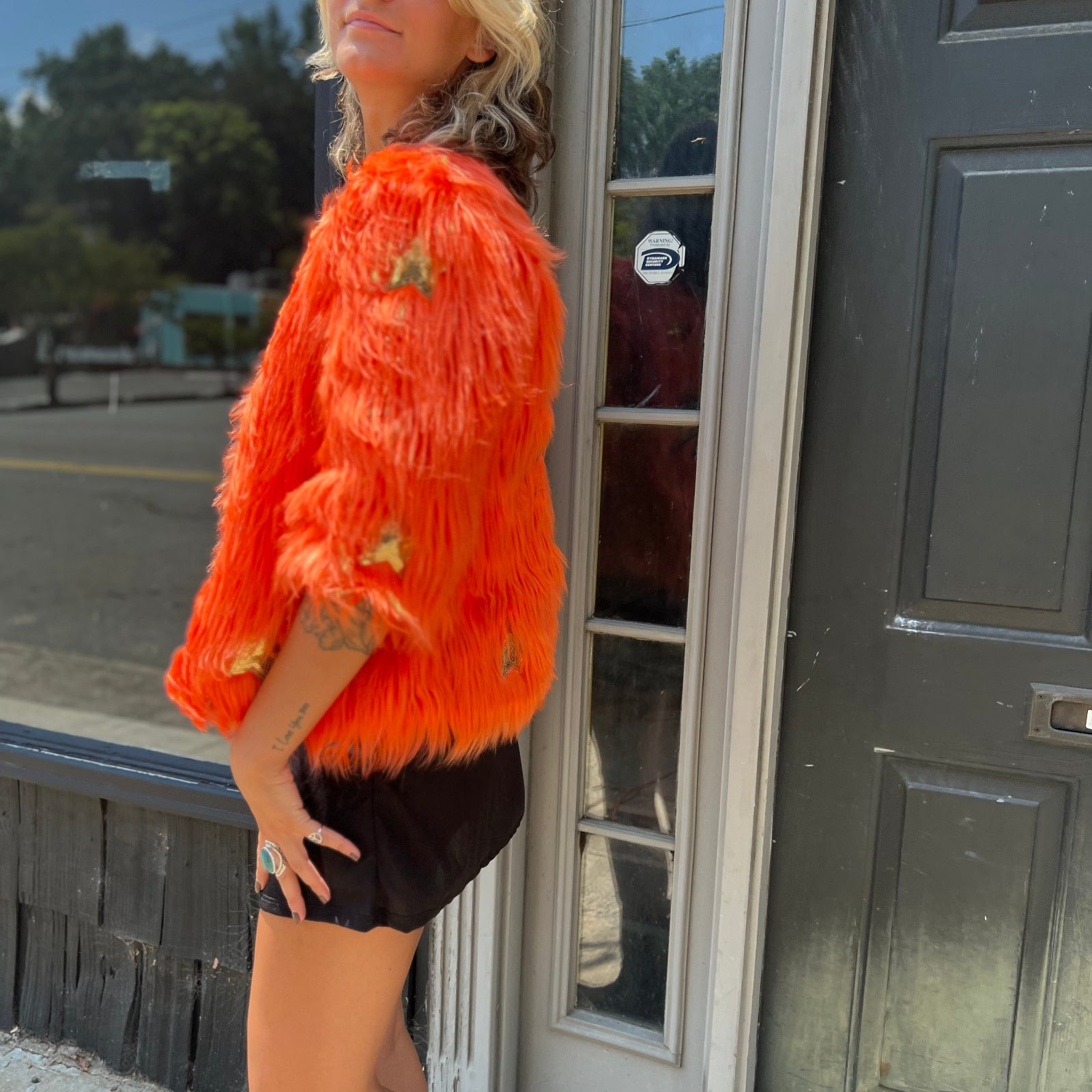 A woman wearing the Planet Queen Orange Faux Fur Jacket with a black dress in front of a window.